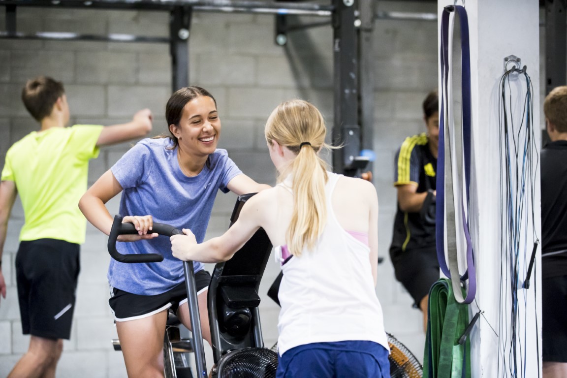 AD Athlete on an exercise bike, smiling at another athlete she's talking to