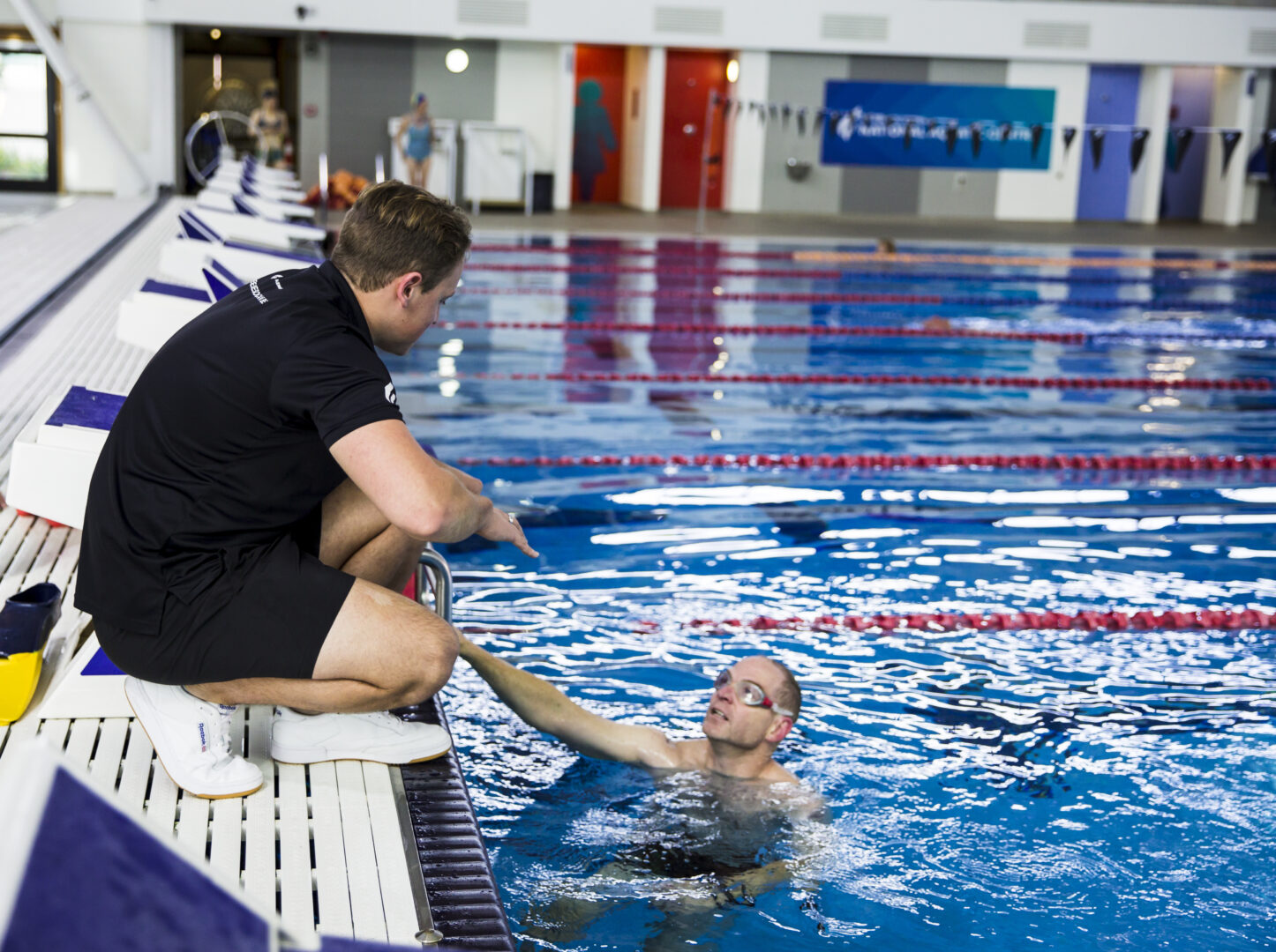 An Adult Swim Squad coach by the dive blocks gives tips to a person in the pool who participates in the Adult Swim Squad class.