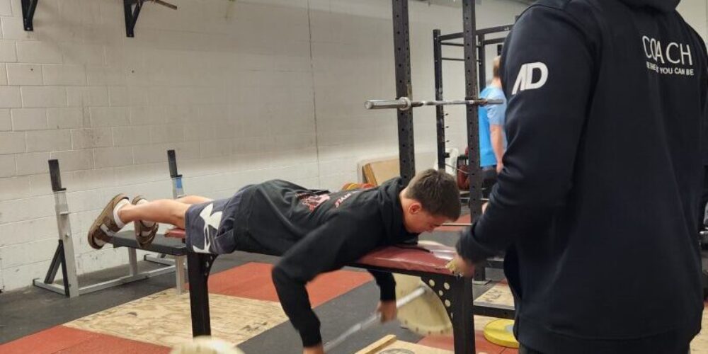 An Athlete Development Coach observes an athlete lifting a barbell from underneath the bench press bench