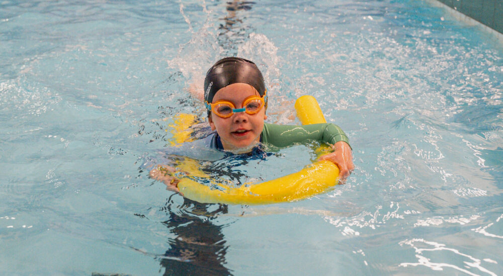 A child in a Swim School practices kicking while holding onto a pool noodle