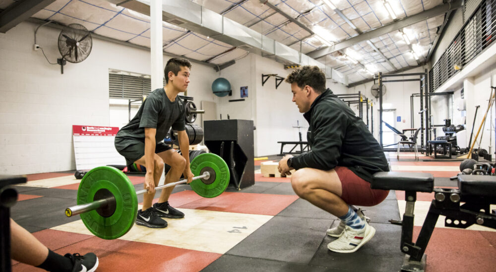 Athlete Development Coach observes an athlete's technique as they lift a weighted barbell