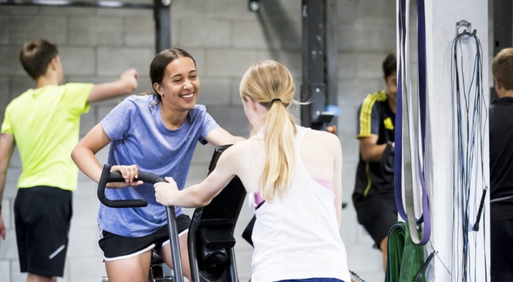 AD Athlete on an exercise bike, smiling at another athlete she's talking to