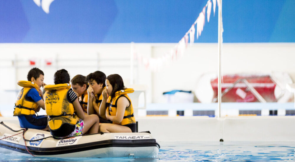 Children with lifejackets on sit in an inflatable boat ready to practice tipping backwards out of the boat