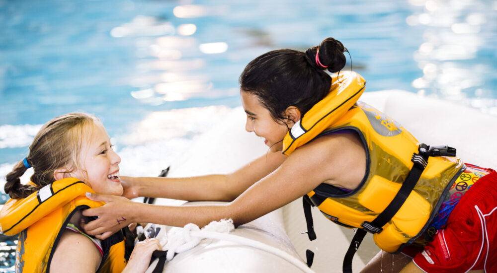 Two children in life jackets, one in a boat and one in the pool