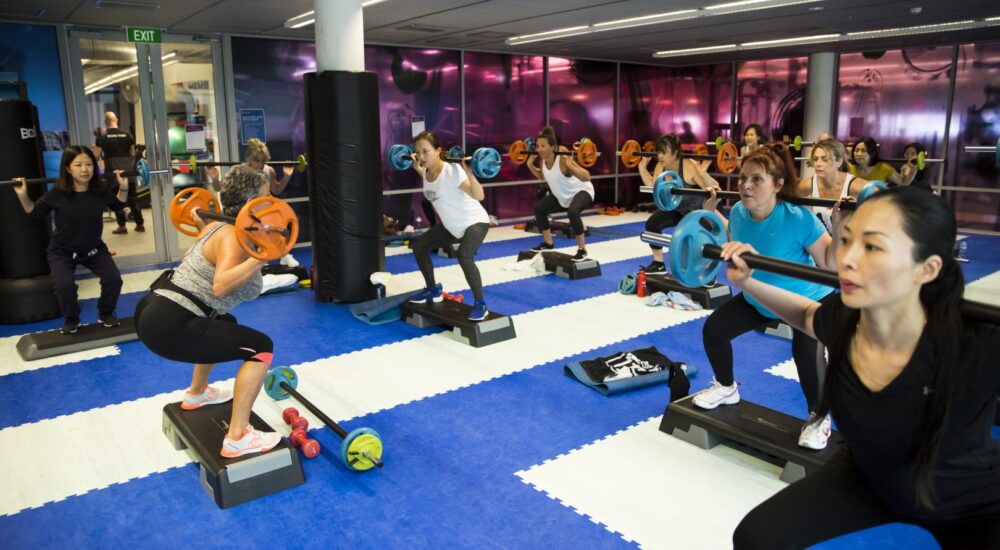 Gym members holding a barbell on their shoulders as they participate in a pump class