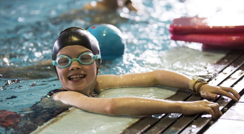 Child in the pool, hanging onto the side as she smiles at the camera